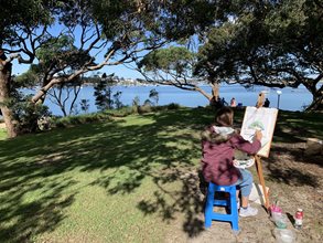 A woman painting on a canvas outside, water in background